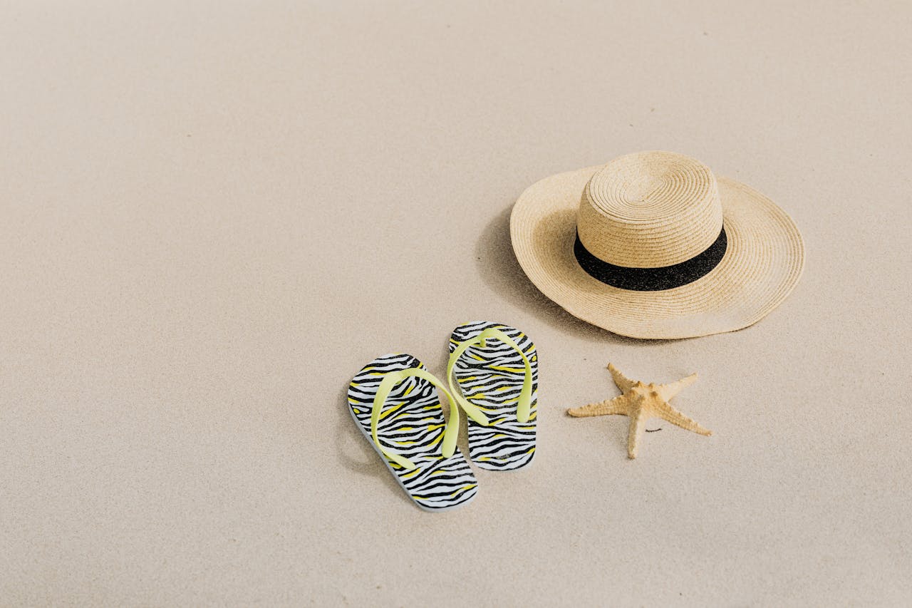 hero-img-01 Flat lay of beach accessories including a straw hat, zebra flip flops, and a starfish on sandy surface.